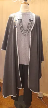 Capa larga gris con vivo blanco en los bordes y botones decorativos. Se cierra con broche en el cuello.