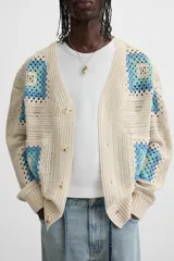 Cárdigan de punto color crudo con diseño de cuadrados de crochet en tonos celeste y azul. Tiene cuello en V, manga larga, bolsillos frontales y cierre con botones.