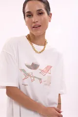 Remera blanca de algodón, corte oversize, con cuello redondo y manga corta. Presenta un estampado frontal con varias reposeras y hamacas de playa, y el texto "LA SIESTA".