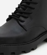 Bota de caña corta para mujer confeccionada en poliuretano (PU) color negro. Presenta punta redonda, suela tipo track con taco mediano de 3,7 cm, cordones frontales con pasadores metálicos y cierre de cremallera lateral.