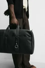 Bolso de viaje negro con compartimento principal con cierre de cremallera, bolsillo interior con cremallera, detalle de arnés en la parte frontal, dos asas de mano y asa de hombro ajustable y extraíble.