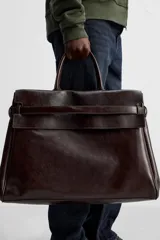Bolso de mano extragrande color marrón oscuro, fabricado en piel de alta calidad. Tiene un compartimento principal con cierre magnético, un bolsillo interior con cremallera y un bolsillo secundario con solapa y cinta. Las asas son de piel.