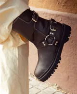 Bota de caña media color marrón oscuro, realizada en cuero encerado. Con hebillas en la caña y herrajes de níquel. El fondo está realizado artesanalmente, es de suela, y es negro. El interior está forrado en forrería vacuna.