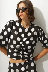Blusa corta negra con estampado de lunares blancos, escote redondo, mangas abullonadas 3/4 y frunce central.