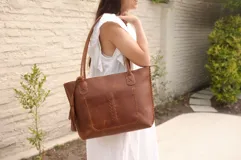 Cartera tote de cuero vacuno color negro, con forrería y cierre. Presenta un bolsillo externo con detalle de costuras y flecos.