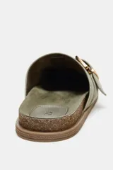 Zueco plano de serraje color verde claro, con tira ajustable y hebilla metálica dorada en la parte delantera. Tiene plantilla aterciopelada, punta redonda y suela de corcho con base de goma.