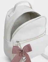 Mochila pequeña de cuero sintético color blanco con bolsillo frontal con solapa y cierre superior. Incluye un charm colgante de moño a rayas rojas y blancas con dije de corazón plateado.