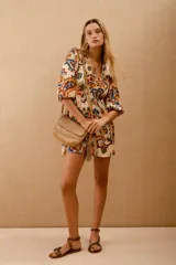 Vestido corto estampado con motivos florales en tonos naranja, azul y verde sobre fondo blanco. Tiene mangas 3/4 con volados en los puños, escote en V y cintura elastizada con lazo.