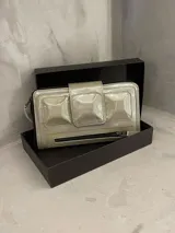 Billetera tipo clutch color beige de cuero charol, con cierre de cremallera y solapa con tres apliques piramidales.