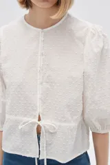 Blusa blanca de algodón con textura de lunares, corte recto, cuello redondo, mangas 3/4 abullonadas y cierre frontal con botones.