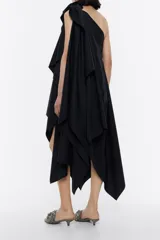 Vestido midi negro asimétrico con un solo hombro y lazo decorativo. Presenta múltiples capas y un corte irregular en el ruedo.