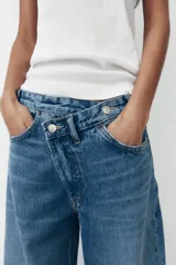 Pantalón de jean celeste de tiro alto, corte wide leg y largo hasta el tobillo. Presenta un diseño asimétrico en la cintura con cierre frontal cruzado con cremallera y botones metálicos.