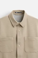 Sobrecamisa negra de tejido con elasticidad, cuello solapa y manga larga acabada en puño con botón a presión. Tiene bolsillos de plastrón en el pecho y cierre frontal con botones a presión.