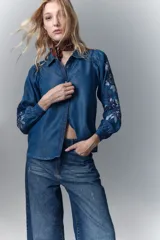 Camisa de calce recto confeccionada en tencel azul denim, con manga larga amplia con frunces en los hombros y cuello camisero. Presenta bordado de hilo en mangas y cartera. Mangas terminadas en puño con botón y ojal. Cartera delantera con botones metálicos con el logo de la marca.