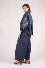 Pantalón ancho de algodón color celeste claro, con bordados de flores blancas en los bajos. Presenta un corte de tiro alto con pliegues frontales y cierre de botón visible.