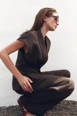 Pantalón blanco de tiro alto, corte ancho y largo cropped, confeccionado en mezcla de lino. Presenta bolsillos laterales, pinzas y raya planchada en delantero. Cierre frontal con cremallera, botón y ganchos metálicos.