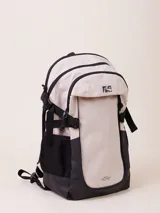 Mochila de nylon ripstop color beige y negro, con compartimento frontal con cierre y logo de Rusty. Cuenta con correas ajustables, bolsillos laterales de malla negra y base reforzada.
