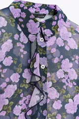 Camisa semitransparente de manga larga con estampado de flores color lila y cuello solapa. Presenta volantes en el frente y cierre con botones.