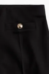 Pantalón negro de corte ancho, tiro alto, con bolsillos traseros decorativos con botón dorado y pliegues delanteros.