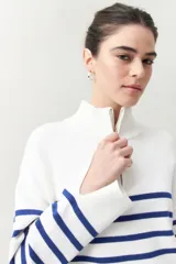 Polera blanca de punto con cuello alto y cierre metálico, de calce relajado y diseño a rayas horizontales azules.