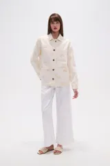 Campera de algodón color crudo, estilo camisa o chaqueta ligera, con bordados en forma de paisley en tono beige claro. Posee cuello camisero, cierre frontal con botones marrones y bolsillos delanteros.