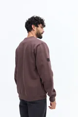 Buzo de algodón color marrón oscuro (ciruela/ciruela quemado) con cuello redondo, puños y ruedo acanalados. Presenta un pequeño bordado de motivos florales en el pecho.