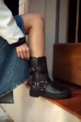 Bota negra de cuero con caña media, suela track, correa con hebilla en el tobillo y arnés con argolla metálica.