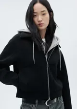 Chaqueta negra corta con capucha gris desmontable y ajustable con cordones. Presenta cuello camisero, cierre de cremallera doble, bolsillos laterales y bajo con goma elástica.