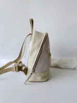 Mochila pequeña de cuero color gris claro y beige, con cierre superior y bolsillo frontal con cierre. Tiene asa de mano y correas ajustables para los hombros.