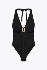 Traje de baño enterizo negro con escote halter atado al cuello y escote en V pronunciado. Presenta un aro metálico dorado en el centro del torso y espalda baja descubierta con banda cruzada.