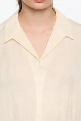 Camisa corta de corte boxy, confeccionada en tejido fantasía texturado color crema o amarillo muy pálido. Presenta cuello camisero, botones frontales y manga 3/4 con sisa ajustable mediante tiras de tejido, además de pespuntes decorativos negros que realzan el borde de las mangas.