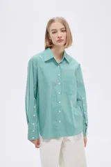 Camisa a rayas verticales verdes y blancas, de corte holgado, manga larga con puños abotonados, cuello clásico y bolsillo de parche en el pecho.