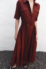 Vestido midi camisero color ladrillo, confeccionado en popelina de algodón. Presenta cuello solapa, manga corta con puños abotonados, bolsillos laterales ocultos y bolsillos de parche con solapa en el pecho. Incluye cinturón lazada en el mismo tejido y cierre frontal con botones ocultos.