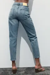 Pantalón de jean gris oscuro, de corte mom fit, tiro alto y largo tobillero. Cuenta con cinco bolsillos, cierre frontal con cremallera y botón.