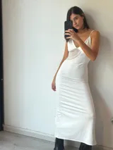 Vestido largo blanco, ajustado al cuerpo, con escote pronunciado en V y tirantes finos. Presenta un detalle de encaje o puntilla en el lateral del busto.