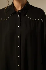 Camisa holgada de manga larga, color negro, con cuello camisa y botones frontales. Presenta apliques de tachas plateadas en los hombros y a lo largo de la tapeta frontal.