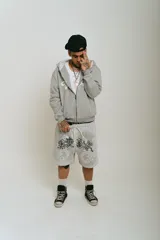 Bermuda de felpa color gris claro con efecto lavado (washed). Presenta un corte ancho con cintura elástica y cordón beige. El diseño frontal incluye un estampado serigrafiado en negro con texto y motivos florales/ornamentales en bordado blanco. Posee remaches metálicos cromados en los laterales y bolsillos.