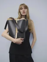 Bolso tipo tote de cuero negro con detalles en beige. Tiene un compartimento principal con cierre magnético, asa larga regulable con hebilla metálica, pespuntes decorativos a contraste, recortes irregulares y logo grabado.