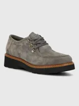 Zapato estilo mocasín o derby de mujer, color gris, confeccionado en gamuza. Presenta suela plataforma negra con borde de cuero marrón cosido y cordones grises que pasan por ojales metálicos dorados.