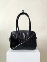 Cartera rígida negra de cuero con tachas plateadas y tres combinaciones de cuero diferentes que aportan textura. Tiene herrajes y cierre metálico plateado marca YKK. Cuenta con un asa fina para llevar al hombro o en la mano y una correa larga extra regulable.