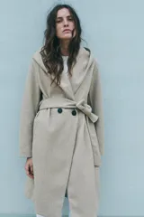 Abrigo largo color beige con capucha, cuello con solapa, manga larga, bolsillos laterales y cinturón lazada en el mismo tejido. Cierre frontal cruzado con botones.