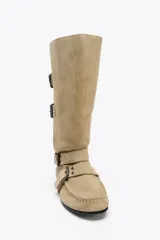 Bota alta color beige de serraje con puntera redondeada. Presenta tres tiras con hebillas metálicas en la parte delantera y caña.