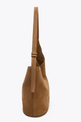 Bolso tipo saca asimétrica color marrón, confeccionado en serraje. Tiene asa bandolera ajustable y cierre con imán. Incluye una cartera con bolsillo extraíble.
