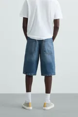Bermuda de denim de algodón, color gris claro lavado, de corte ancho (baggy fit) y largo hasta la rodilla. Presenta cinco bolsillos y cierre frontal con cremallera y botón.