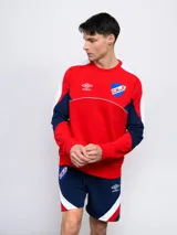 Buzo de felpa azul marino, con mangas largas, cuello redondo y puños acanalados. Presenta cortes en blanco y rojo en la parte superior, logo de Umbro bordado en el pecho y escudo del Club Nacional de Football.