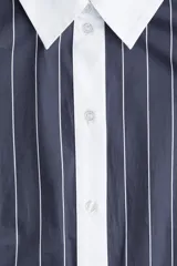 Vestido corto estilo camisero a rayas verticales azul marino y blancas, con cuello y puños blancos. Tiene cierre frontal con botones y lazo para atar en la cintura.