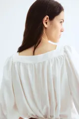 Blusa corta blanca de tejido liviano con escote cuadrado fruncido, corte holgado, manga globo raglán al codo y elástico fino en los puños.