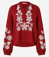 Blusa color borgoña de batista con cuello en V y botones. Presenta bordados florales blancos en el frente y las mangas largas con puños elásticos.