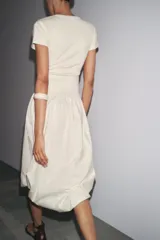 Vestido largo color crema, con cuello redondo y mangas cortas. Presenta corte a la altura de la cintura y falda amplia con terminación redondeada tipo globo.