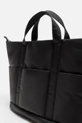 Bolso tipo shopper negro fabricado en tejido técnico. Cuenta con un compartimento principal con cierre de cremallera y un bolsillo interior con cierre. En la parte frontal tiene tres bolsillos. Tiene dos asas de mano.